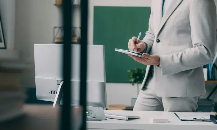 Man in a light suit standing at a modern office desk, writing in a notebook near a computer during work